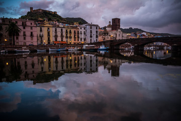 Night city view of Bosa, Sardinia, Italy. Colorful houses and beautiful clouds reflecting in the water after rain at dusk.
