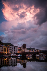 Fototapeta na wymiar Night city view of Bosa, Sardinia, Italy. Colorful houses and beautiful clouds reflecting in the water after rain at dusk.