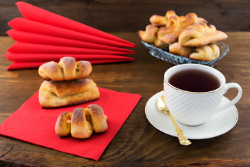 Serving Breakfast-curly pastry with filling, tea in a white porcelain mug, red napkins on a wooden background.