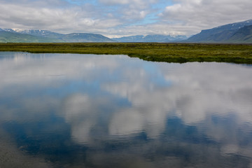 Beautiful cloudy sky reflecting in calm water with fresh green field and mountains in Iceland