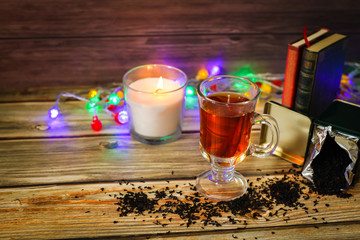 Cozy winter still life: cup of hot tea and books, candle on wooden background and blurred garland lights