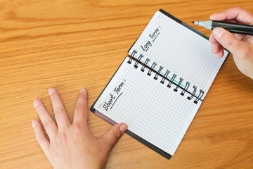 Woman fills in her goals and plans for long and short terms in spiral notepad on wooden table, top view