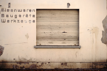 Geschlossenes Eisenwarengeschäft  / Die vergilbte Fassade eines geschlossenen Eisenwarengeschäftes mit heruntergelassenen Rollläden. .