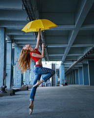 Obraz premium Ballerina is dancing with a yellow umbrella in jeans, T-shirt and pointe. Street performance. Modern ballet. Slim girl dancing on tiptoe. Red hair fly.