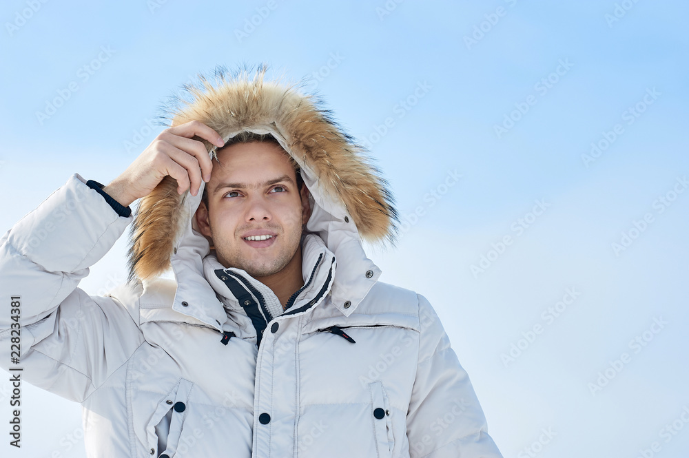 Wall mural Fashion young beautiful man in a white jacket with fur hood in winter