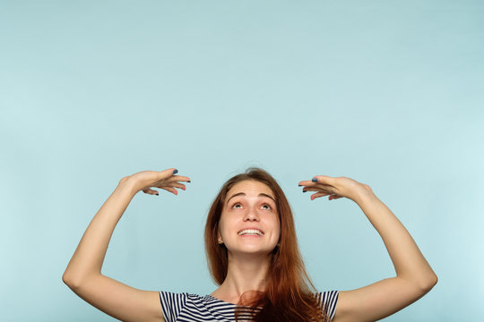 Happy Joyful Smiling Woman Pretending To Hold Smth Above Her Head With Hands. Virtual Message Or Object Pressing Down On Young Beautiful Brown Haired Girl. Advertising Space On Blue Background.