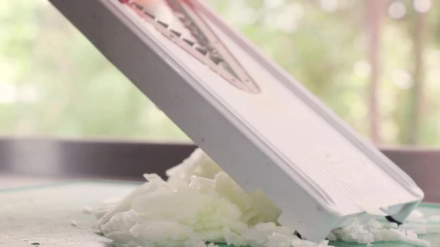 A woman using a mandoline slicer, chopping onions, next to a window on a clean, bright, stainless steel kitchen