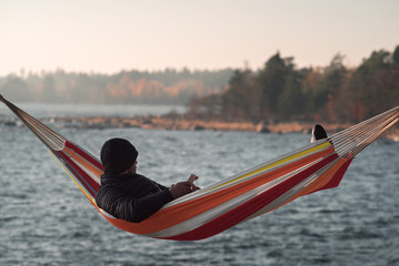 A man sits in a hammock near the lake and admires the autumn forest. Smarfon in his hands.