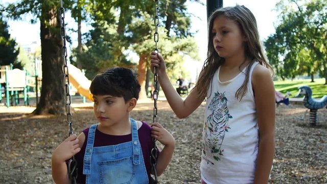 Two girls look serious as they listen to someone on swing-set out of frame