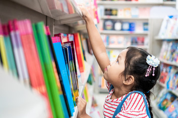 Cute asian child girl select book on bookshelf in bookstore in supermarket - obrazy, fototapety, plakaty
