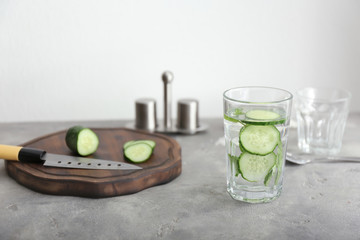 Glass of tasty fresh cucumber water with cutting board on grey table