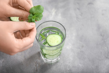 Woman putting mint into glass with cucumber water on grey table, closeup