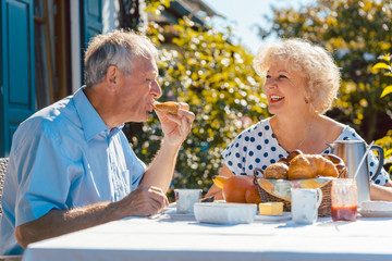 Senior woman and man having breakfast sitting in their garden outdoors in summer, eating bread...