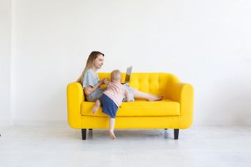 Casual young mom designer sitting on yellow leather couch indoors white room with laptop pc on her lap, using internet for remote work, looking smiling as her little son came to distract her from work