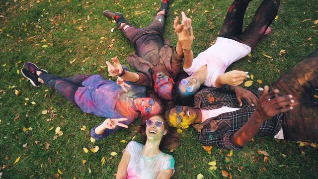 Slow motion portrait of relaxed people lying on grass in park with coloured faces and clothing, looking at camera and smiling moving hands. Party and nature concept.