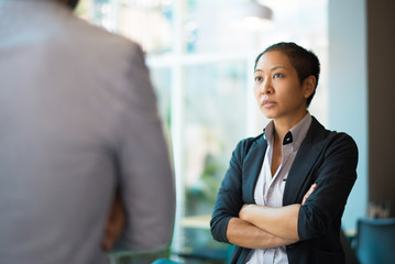 Tense Asian businesswoman looking at male partner with crossed arms. Two colleagues confronting each other in office space. Clashing personalities concept