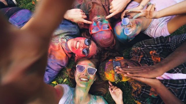 Point of view shot of happy men and women with painted faces lying on grass at Holi festival and taking selfie posing for camera. People are wearing sunglasses and dirty clothing.