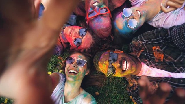Point of view shot of happy woman lying on grass with friends at party Holi festival and recording video taking selfie holding device with camera. Fun and friendship concept.