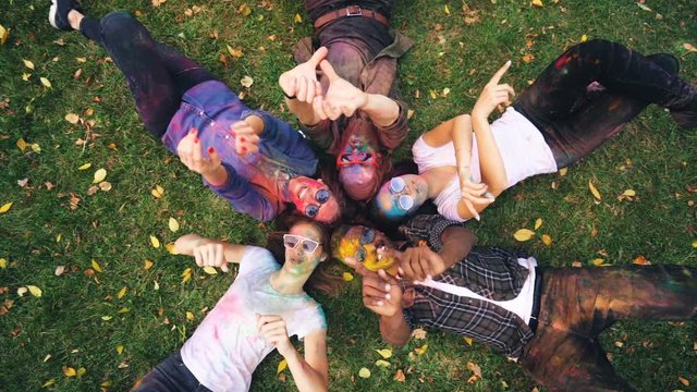 Zoom-in of relaxed people multi-ethnic group lying on grass together with coloured faces and clothes, looking at camera, smiling and moving hands. Relaxation and party concept.