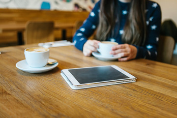 Cappuccino coffee in a mug on the table next to the tablet. The girl is drinking coffee in the background. Relaxing or meeting in a cafe. The girl is waiting for someone else.