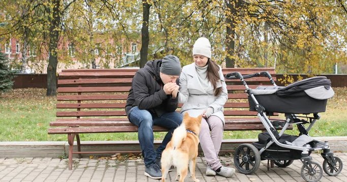 Happy family walking in autumn park with stroller. Mom, Dad and their newborn baby son sit on a bench and play with their family dog