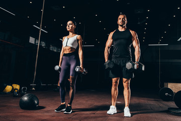fit sportsman and sportswoman exercising with dumbbells together in dark gym