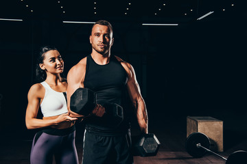 athletic bodybuilder exercising with barbell and looking at camera while woman standing behind in dark gym