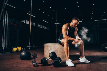 handsome athletic man sitting on cube with gym equipment around and clapping hands with talc before workout