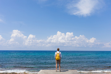 Tourist traveler guy stands on a wooden pier in solitude and looks at the blue sea, ocean. Calm and peace. Yellow backpack. The coast and a sandy beach with stones. Travel and vacation with a hike.