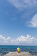 Tourist traveler guy stands on a wooden pier in solitude and looks at the blue sea, ocean. Calm and peace. Yellow backpack. The coast and a sandy beach with stones. Travel and vacation with a hike.