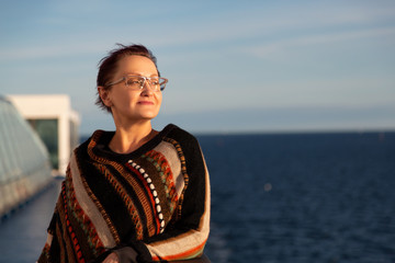 Nice portrait of an older woman looking out to the sea and cruising on a cruise ship. Beautiful middle aged lady wearing colorful jumper /poncho and standing on a cruise ship deck on sunny day. - 228271173