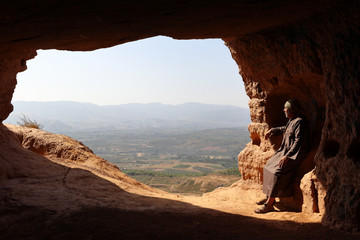ISOLATED HERMIT SITTING AT THE ENTRANCE OF A CAVE ON THE HIGH OF A MOUNTAIN