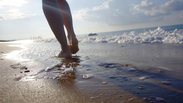 tilt up view of slim legs of young woman walking on seashore, waves are rolling to coast