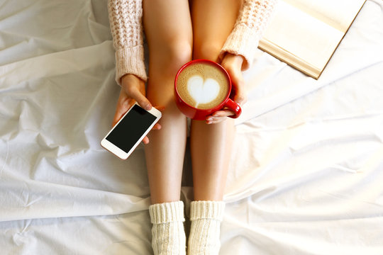 Lazy afternoon concept. Close up shot of woman sitting in bed, reading book, holding red cup of cappuccino coffee with heart shaped latte art foam & blank screen mobile phone. Background, copy space.