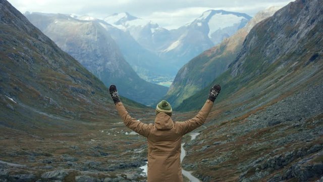 Female traveler looking down from top of the mountain to epic landscape view to valley and fjord in Norway and raising her hands