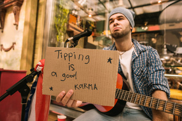 Good karma. Dissatisfied male musician holding cardboard with inscription and seating