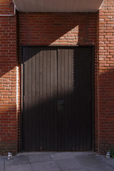 Sunlit red brick wall with dark wooden double leaved door and diagonal hard shadow