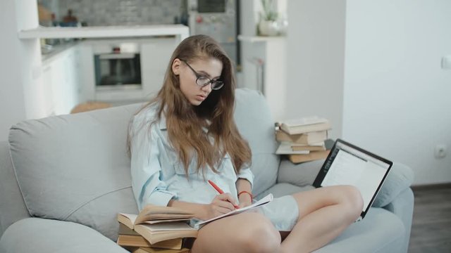 Pretty teenage girl in glasses with a puzzled look is reading a text on a laptop, looking at screen, write something in notebook, preparing for exams or homework, lot of books