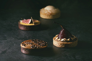 Variety of sweet tartlets with chocolate, caramel, pears, figs on black texture table.