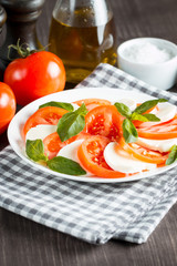 Photo of Caprese Salad with tomatoes, basil, mozzarella, olives and olive oil on wooden background. Italian traditional caprese salad ingredients. Mediterranean, organic and natural food concept.