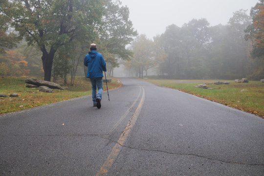 Lone man in blue jacket and pole walks alone down the road on a foggy morning in the fall
