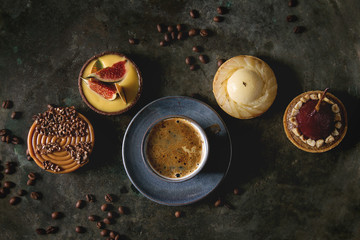 Variety of sweet tartlets with chocolate, caramel, pears, figs with cup of coffee and coffee beans around on black texture background. Flat lay, space