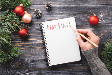 Mockup with white paper notebook and Christmas decorations on wooden table. Woman's hand writing letter for Santa in notepad. Top view.