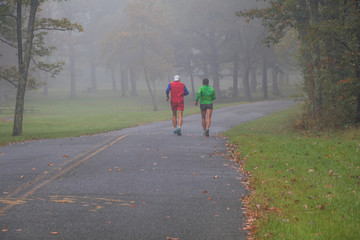 Pair of fit male mature runners in bright clothing through a bleak foggy morning in the fall; dedication to fit lifestyle concept 