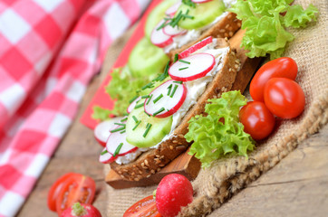 Close-up of wholemeal grain bread with curd cheese, fresh radish, cucumber and tomatoes on a wooden cutting board - concept of healthy fitness breakfast or snack