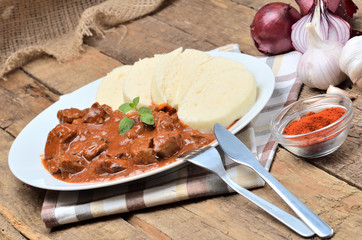 Pork goulash meat with dumplings on white plate, cutlery, garlic, onion, pepper, tablecloth in the background - typical Czech food