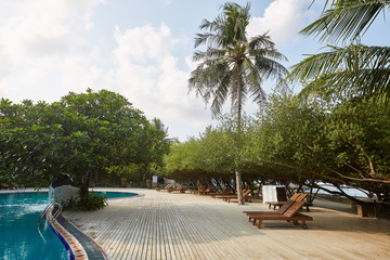 Swimming pool side of luxury hotel ith deck chairs, palm trees and blue ocean. Maldives