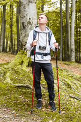 Fototapeta premium Young blonde attractive man with trekking poles standing in the forest, he searching some places and enjoying nature, trekking, recreation and healthy lifestyle concept