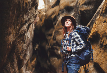 Travel young woman wearing brown hat, plaid shirt, jeans and brown boots with backpack walking in canyon with moss on rocks after hiking, travelling concept