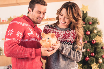 smiling couple holding piggy bank with savings at home with christmas tree